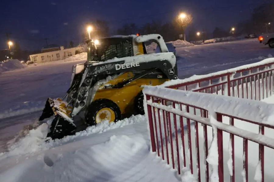Snow-covered train platform at a transport station in Manhasset, New York, with a vehicle clearing snow from the road during a major winter storm.